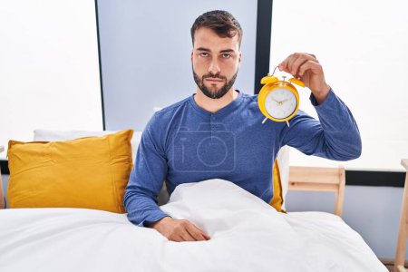 Photo for Handsome hispanic man in the bed holding alarm clock thinking attitude and sober expression looking self confident - Royalty Free Image