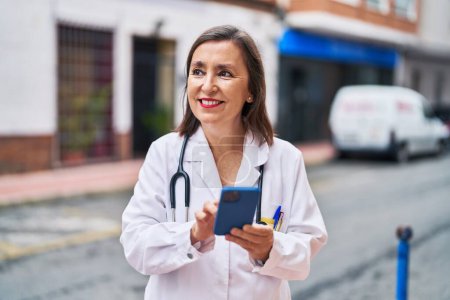 Photo for Middle age woman wearing doctor uniform using smartphone at street - Royalty Free Image