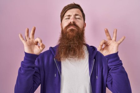 Foto de Hombre caucásico con barba larga de pie sobre fondo rosa relajarse y sonreír con los ojos cerrados haciendo gesto de meditación con los dedos. concepto de yoga. - Imagen libre de derechos