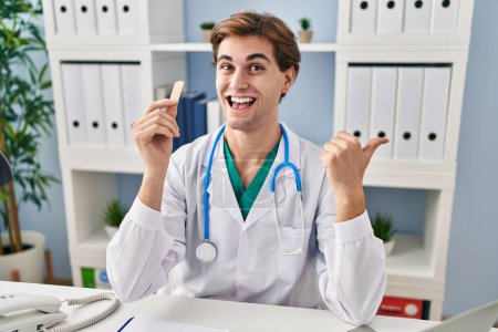 Photo for Young doctor man holding band aid pointing thumb up to the side smiling happy with open mouth - Royalty Free Image