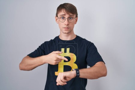 Téléchargez les photos : Homme blond caucasien portant Bitcoin t-shirt à la hâte pointant pour regarder le temps, l'impatience, bouleversé et en colère pour délai - en image libre de droit