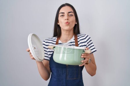 Photo for Young brunette woman wearing apron holding cooking pot looking at the camera blowing a kiss being lovely and sexy. love expression. - Royalty Free Image