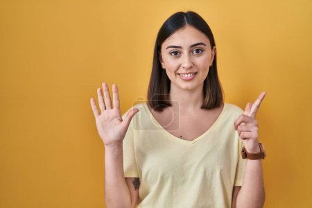 Foto de Chica hispana vistiendo camiseta casual sobre fondo amarillo mostrando y señalando con los dedos número siete mientras sonríe confiado y feliz. - Imagen libre de derechos