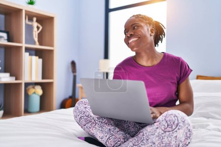 Téléchargez les photos : Femme afro-américaine utilisant un ordinateur portable assis sur le lit dans la chambre - en image libre de droit