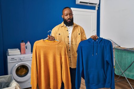 Photo for African american man holding clean clothes on hangers at laundry room skeptic and nervous, frowning upset because of problem. negative person. - Royalty Free Image