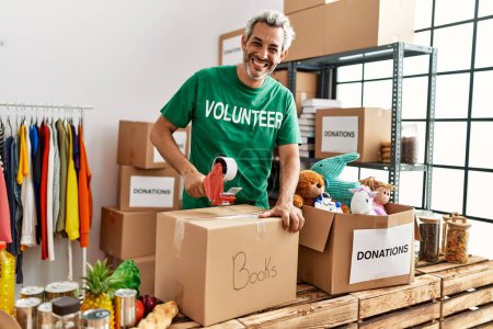 Foto de Hombre de pelo gris de mediana edad voluntario sonriendo confiado embalaje libros caja de cartón en el centro de caridad - Imagen libre de derechos