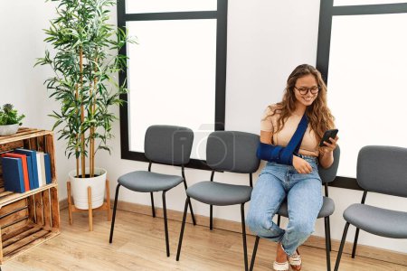 Téléchargez les photos : Jeune belle femme hispanique blessée assise sur une chaise en utilisant un smartphone dans la salle d'attente de la clinique - en image libre de droit