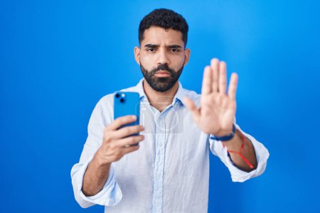 Photo for Hispanic man with beard using smartphone typing message doing stop sing with palm of the hand. warning expression with negative and serious gesture on the face. - Royalty Free Image