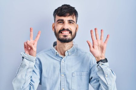 Foto de Joven hombre hispano con barba de pie sobre fondo azul mostrando y señalando hacia arriba con los dedos número siete mientras sonríe confiado y feliz. - Imagen libre de derechos