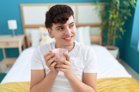 Téléchargez les photos : Homme non binaire buvant une tasse de café assis sur le lit dans la chambre - en image libre de droit
