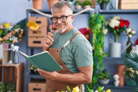 Foto de Florista hombre de pelo gris de mediana edad sonriendo confiado libro de lectura en la tienda de flores - Imagen libre de derechos
