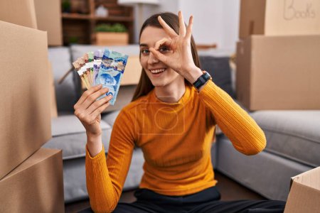 Foto de Joven mujer caucásica sentada en el suelo en casa nueva sosteniendo el dinero canadiense sonriendo feliz haciendo señal de ok con la mano en el ojo mirando a través de los dedos - Imagen libre de derechos