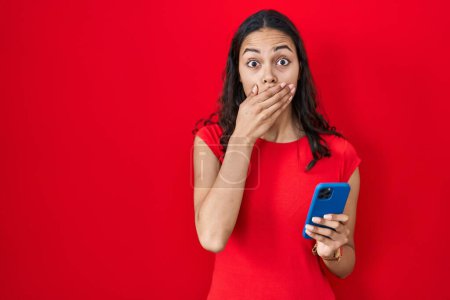 Téléchargez les photos : Jeune femme brésilienne utilisant smartphone sur fond rouge choqué couvrant la bouche avec les mains pour erreur. concept secret. - en image libre de droit
