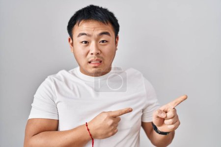 Téléchargez les photos : Jeune homme chinois debout sur fond blanc pointant de côté inquiet et nerveux avec les deux mains, préoccupé et expression surprise - en image libre de droit