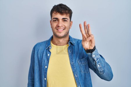 Photo for Young hispanic man standing over blue background showing and pointing up with fingers number three while smiling confident and happy. - Royalty Free Image