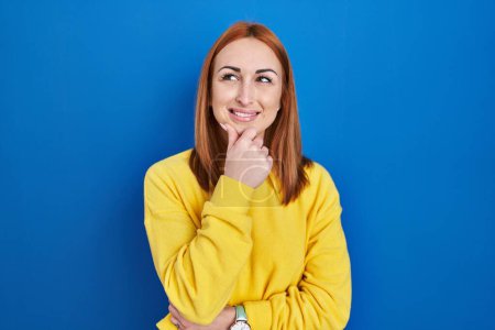 Photo for Young woman standing over blue background looking confident at the camera smiling with crossed arms and hand raised on chin. thinking positive. - Royalty Free Image