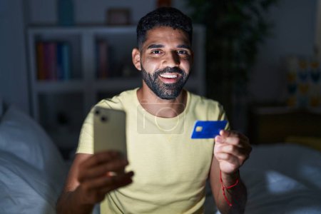 Téléchargez les photos : Jeune homme arabe utilisant ordinateur portable et carte de crédit assis sur le lit dans la chambre - en image libre de droit