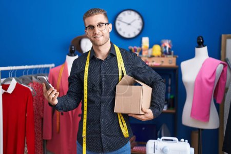 Photo for Young caucasian man tailor using smartphone holding delivery package at clothing factory - Royalty Free Image