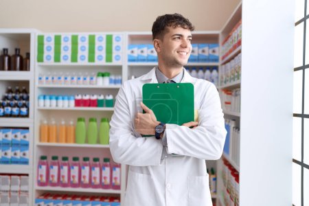 Foto de Joven farmacéutico hispano sonriendo confiado sosteniendo portapapeles en la farmacia - Imagen libre de derechos