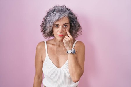 Téléchargez les photos : Middle age woman with grey hair standing over pink background pointing to the eye watching you gesture, suspicious expression - en image libre de droit