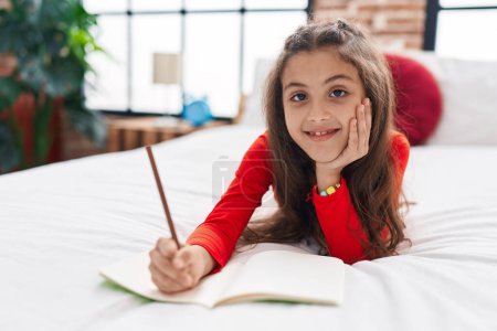 Photo for Adorable hispanic girl writing on notebook lying on bed at bedroom - Royalty Free Image