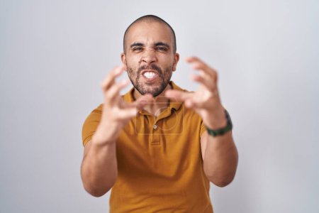 Foto de Hombre hispano con barba de pie sobre fondo blanco gritando frustrado por la rabia, manos tratando de estrangular, gritando loco - Imagen libre de derechos