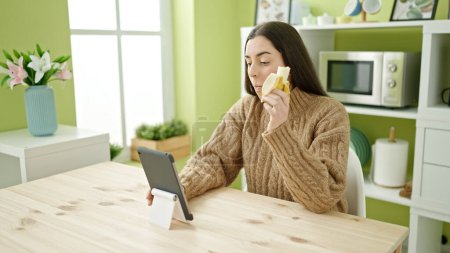 Photo for Young beautiful hispanic woman eating banana watching video on touchpad at kitchen - Royalty Free Image