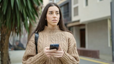 Téléchargez les photos : Jeune belle femme hispanique utilisant un smartphone avec une expression sérieuse dans la rue - en image libre de droit