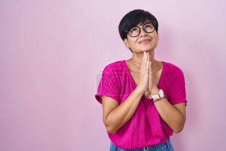 Foto de Joven mujer asiática con el pelo corto de pie sobre fondo rosa mendigando y rezando con las manos junto con la esperanza de expresión en la cara muy emocional y preocupado. mendicidad. - Imagen libre de derechos