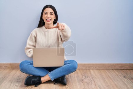 Téléchargez les photos : Jeune femme utilisant un ordinateur portable assis sur le sol à la maison en riant de vous, pointant du doigt la caméra avec la main sur le corps, expression de honte - en image libre de droit