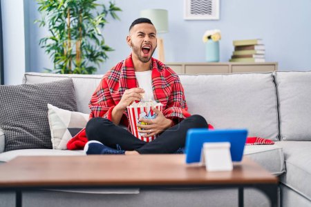 Photo for Young hispanic man eating popcorn watching movie on tablet device angry and mad screaming frustrated and furious, shouting with anger looking up. - Royalty Free Image