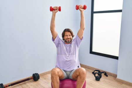 Photo for Middle age man training with dumbbells sitting on fit ball at sport center - Royalty Free Image