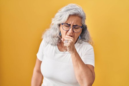 Téléchargez les photos : Femme d'âge moyen avec les cheveux gris debout sur fond jaune se sentant mal à l'aise et la toux comme symptôme de rhume ou de bronchite. concept de soins de santé. - en image libre de droit