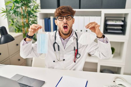 Photo for Arab man with beard wearing doctor uniform holding medical mask angry and mad screaming frustrated and furious, shouting with anger looking up. - Royalty Free Image