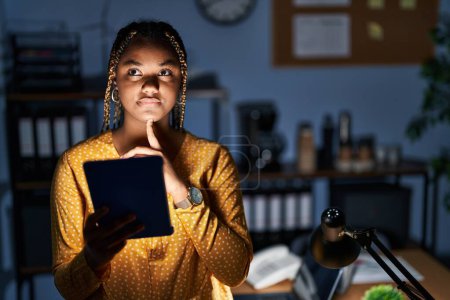 Photo for African american woman with braids working at the office at night with tablet thinking concentrated about doubt with finger on chin and looking up wondering - Royalty Free Image