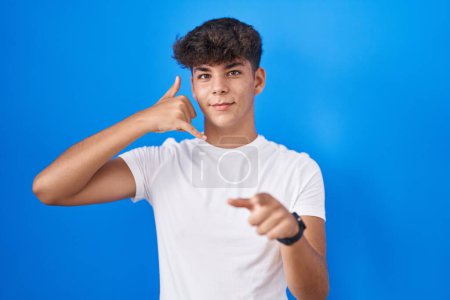 Téléchargez les photos : Adolescent hispanique debout sur fond bleu souriant faire parler au téléphone geste et vous pointant du doigt. Appelle-moi. - en image libre de droit