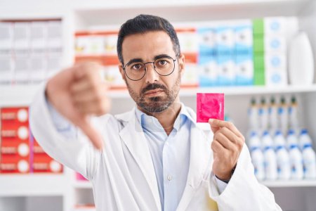 Téléchargez les photos : Homme hispanique avec barbe travaillant à la pharmacie pharmacie tenant un préservatif avec un visage en colère, signe négatif montrant aversion avec les pouces baissés, concept de rejet - en image libre de droit