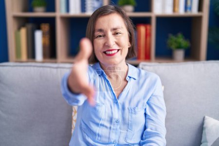 Foto de Mujer hispana de mediana edad sentada en el sofá en casa sonriendo amigable ofreciendo apretón de manos como saludo y bienvenida. negocios exitosos. - Imagen libre de derechos