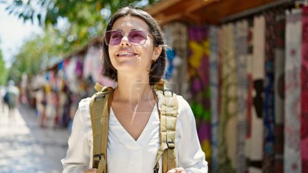 Photo for Young beautiful hispanic woman tourist smiling confident wearing backpack at street market - Royalty Free Image