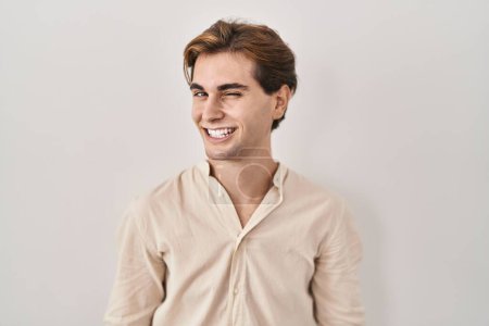 Foto de Young man standing over isolated background winking looking at the camera with sexy expression, cheerful and happy face. - Imagen libre de derechos