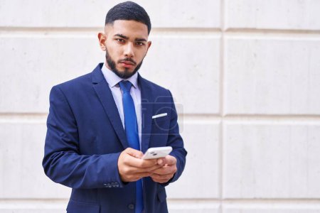 Téléchargez les photos : Jeune homme d'affaires latino-américain utilisant un smartphone avec une expression sérieuse sur fond blanc isolé - en image libre de droit