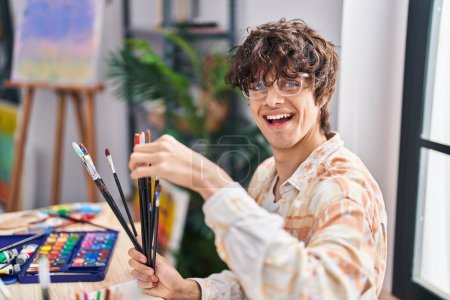 Téléchargez les photos : Jeune homme hispanique tenant des pinceaux assis sur le bureau au studio d'art - en image libre de droit