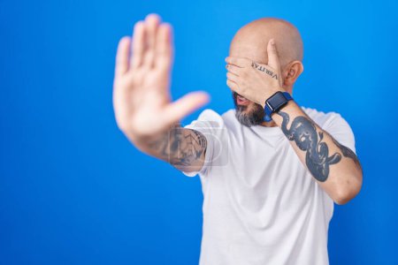 Foto de Hombre hispano con tatuajes de pie sobre fondo azul cubriendo los ojos con las manos y haciendo un gesto de stop con expresión triste y de miedo. concepto avergonzado y negativo. - Imagen libre de derechos