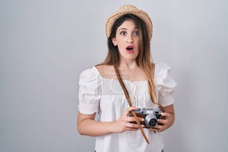Téléchargez les photos : Jeune femme brune tenant une caméra vintage en état de choc, à l'air sceptique et sarcastique, surprise avec la bouche ouverte - en image libre de droit
