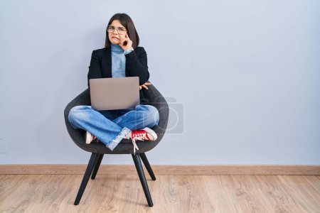 Foto de Mujer hispana joven sentada en silla usando computadora portátil pensando preocupado por una pregunta, preocupado y nervioso con la mano en la barbilla - Imagen libre de derechos