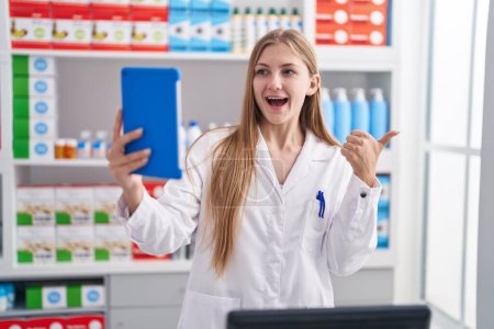 Photo for Young caucasian woman working at pharmacy drugstore doing video call with tablet pointing thumb up to the side smiling happy with open mouth - Royalty Free Image