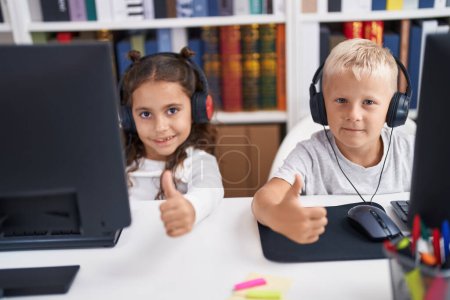 Téléchargez les photos : Adorable garçon et fille élèves utilisant l'ordinateur et écouteurs faisant pouce vers le haut geste à la salle de classe - en image libre de droit