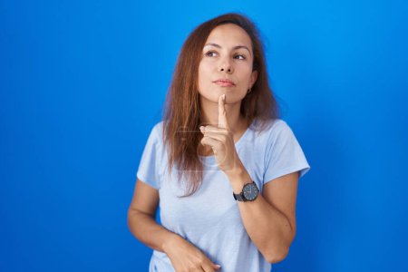 Téléchargez les photos : Femme brune debout sur fond bleu pensant concentré sur le doute avec le doigt sur le menton et regardant vers le haut se demandant - en image libre de droit
