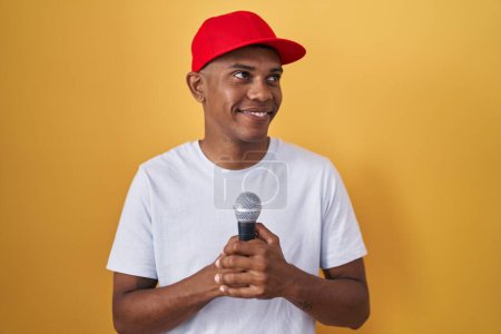 Téléchargez les photos : Jeune homme hispanique chantant chanson en utilisant le microphone souriant regardant vers le côté et regardant loin en pensant. - en image libre de droit