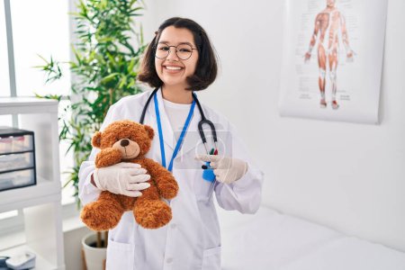 Foto de Joven doctora hispana sosteniendo oso de peluche sonriendo feliz señalando con la mano y el dedo - Imagen libre de derechos